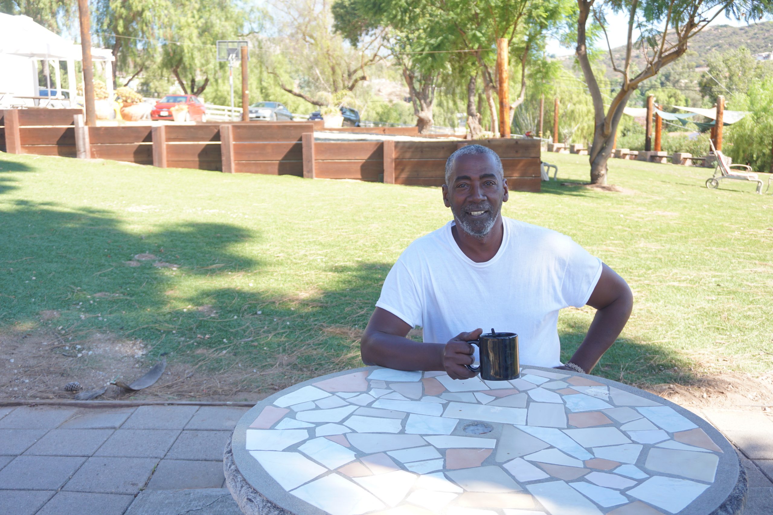 A man in a white shirt, sitting outside in the shade at a table smiling and holding a coffee cup. He is a part of what makes veteran housing communities important and successful.