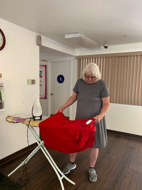 An older woman with white hair and glasses, wearing a grey dress ironing a red shirt on an ironing board at Cambridge Gardens senior housing community.