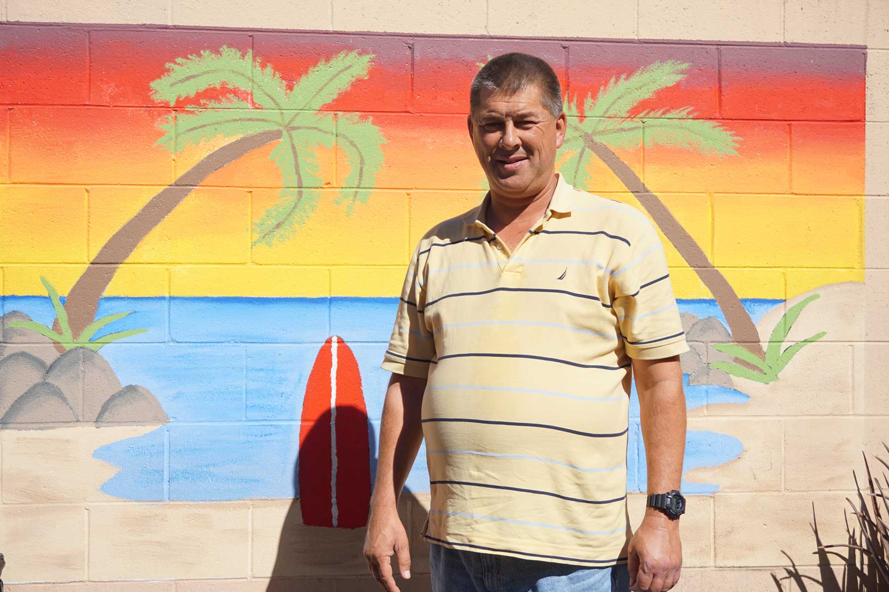 A man who is a suportive housing resident of the Luhman Center for Supportive Living adult residential care facility stands in front of a wall mural depicting a beach sunset, including sand, water, two palm trees and a red surfboard.