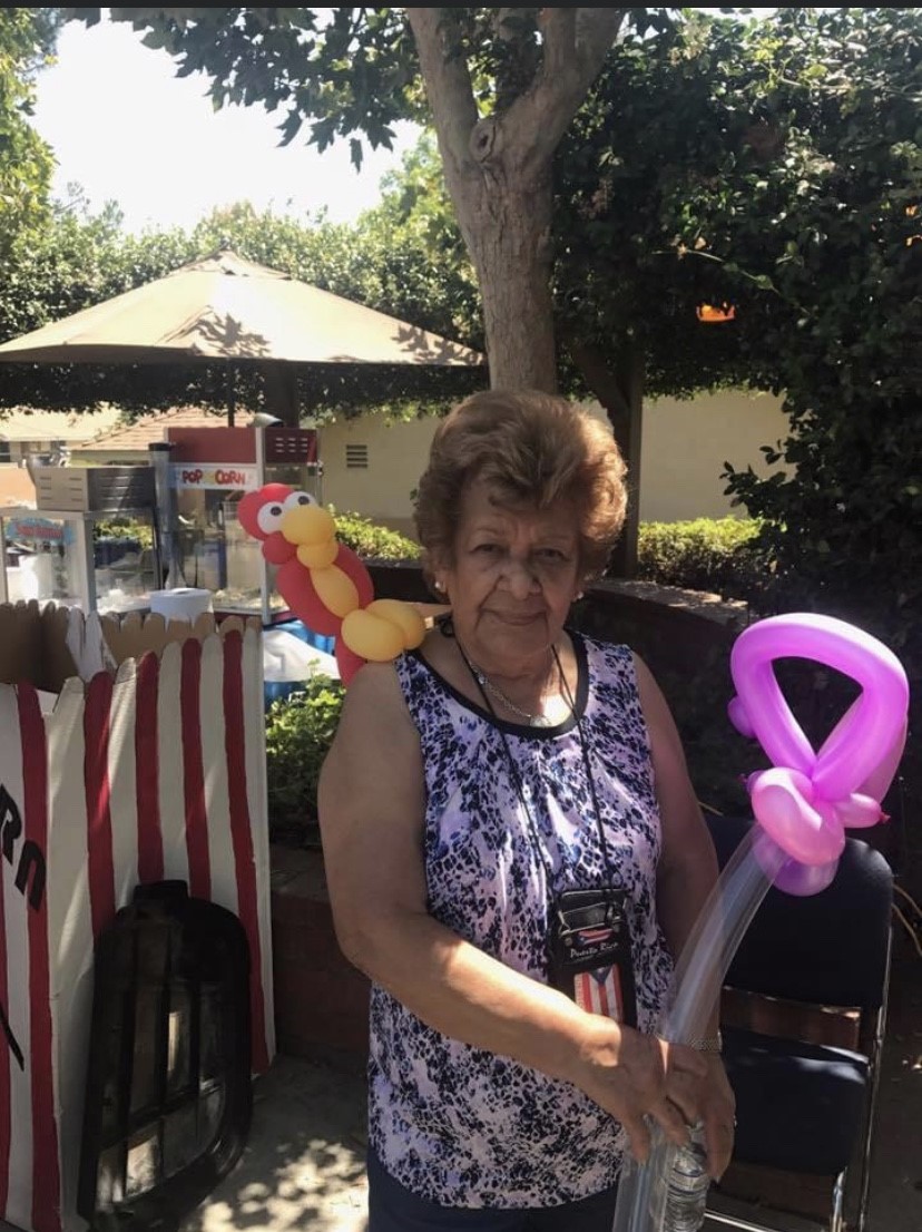 An older woman outside holding a purple balloon sculpture with a yellow and red balloon parrot on her shoulder at Cambridge Gardens senior housing community.