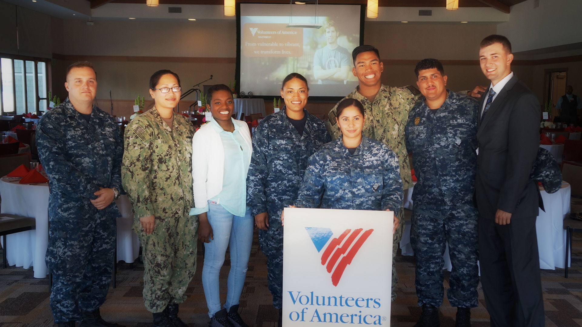 A photo of six people standing, five in military uniform, smiling and holding a VOASW banner, symbolic of Volunteers of America Southwest contact info.
