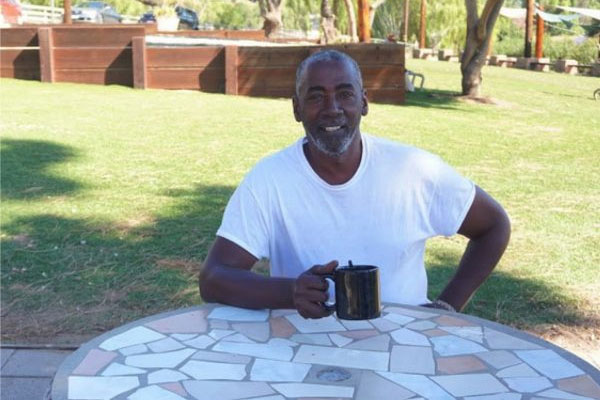 An older man wearing a white shirt, sitting with a coffee cup at a table in the shade at a supportive housing facility.