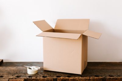 Photo of an empty, open cardboard moving box and roll of packing tape sitting on a dark reclaimed wood console table set against a white wall, symbolizing the need for rental assistance in san diego.