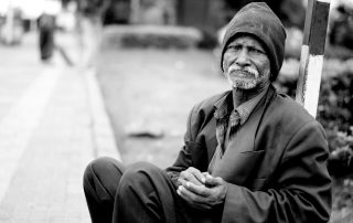 A black and white photo of an African-American man wearing a beanie, overcoat and pants while clasping is hands as he sits on a curb in the city. The image is meant to hightlight coronavirus among San Diego homeless population.