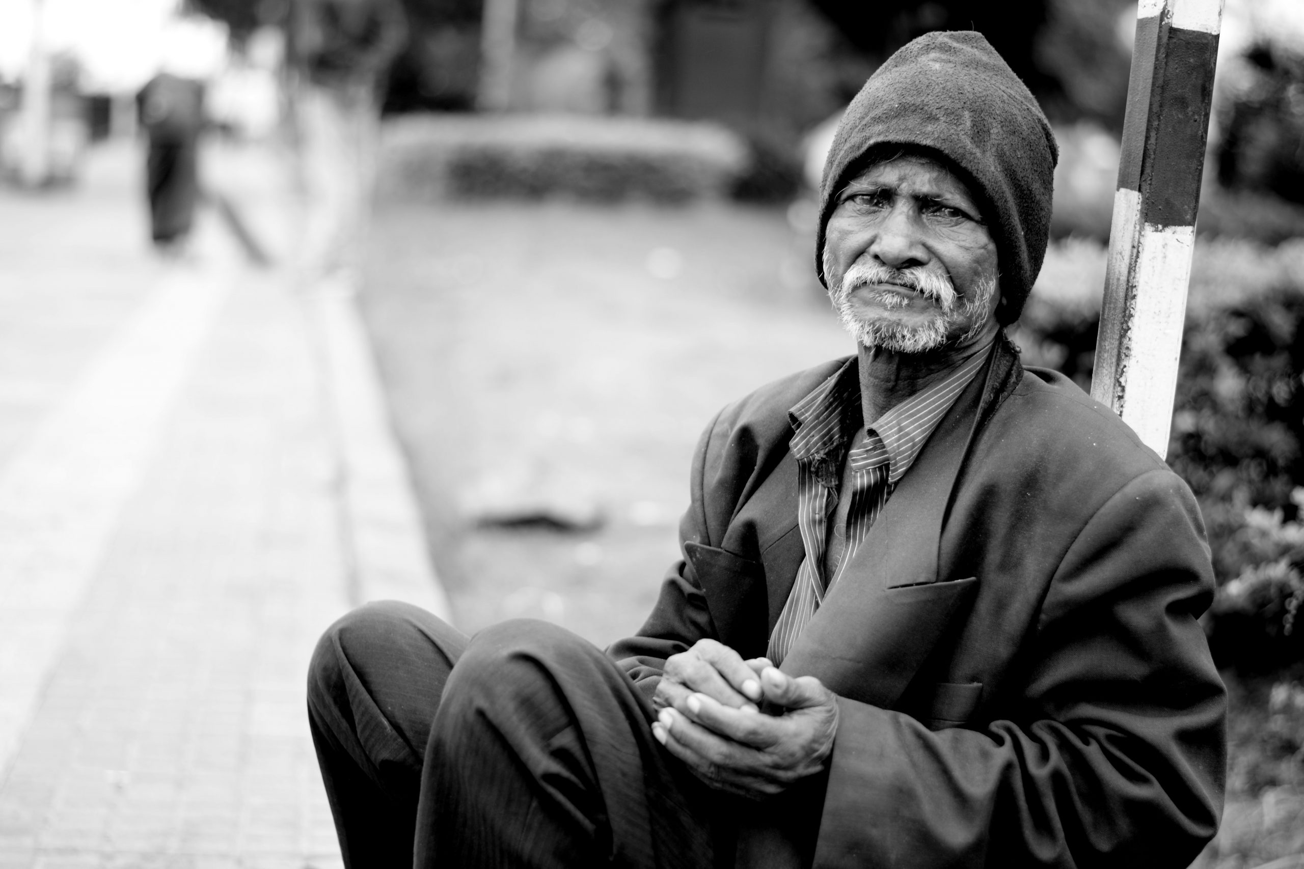 A black and white photo of an African-American man wearing a beanie, overcoat and pants while clasping is hands as he sits on a curb in the city. The image is meant to hightlight coronavirus among San Diego homeless population.