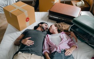 Photo of a couple laying on a bed and looking into each other's eyes, surrounded by packing boxes and two suitcases, meant to symbolize symbolizing the need for rental assistance in San Diego.