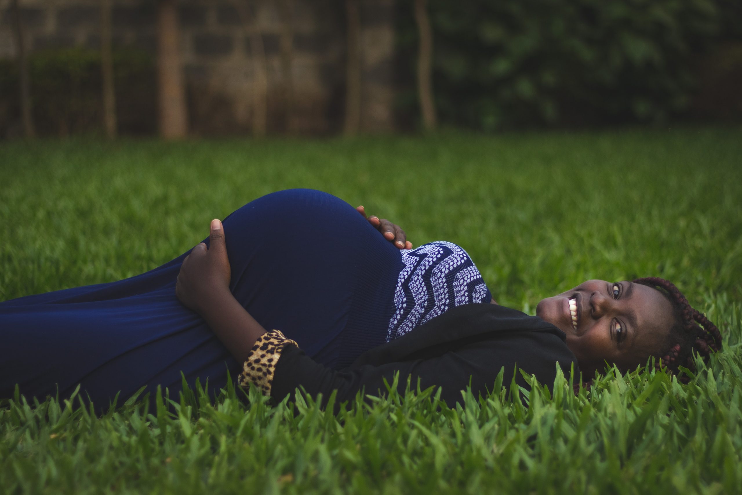 A smiling, pregnant Black women wearing a blue dress lays in green grass, symbolizing the risks of being pregnant with coronavirus.
